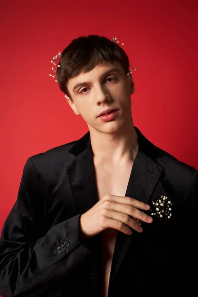 Bel homme en tenue élégante avec des fleurs dans les cheveux en regardant la caméra sur fond rouge — Photo de stock