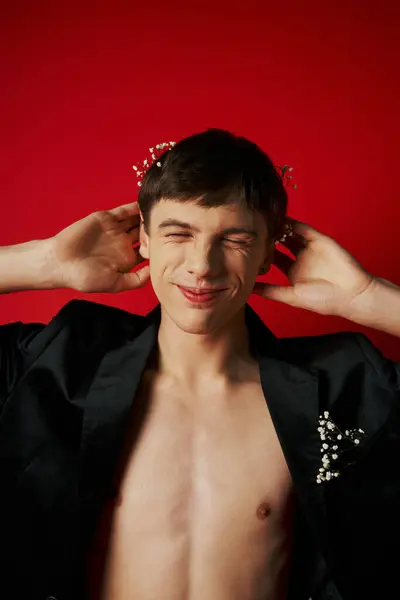 Homem feliz em blazer de veludo elegante com flores no cabelo sorrindo com olhos fechados no fundo vermelho — Fotografia de Stock