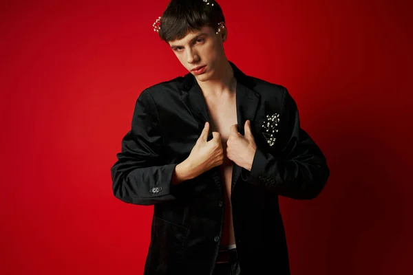 Elegant young man in velvet blazer with flowers in hair looking at camera and posing on red backdrop — Stock Photo