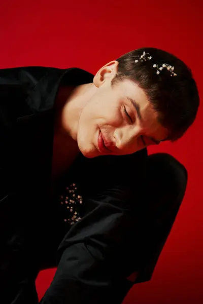 Joyeux jeune homme en velours noir veste avec des fleurs dans les cheveux souriant sur fond rouge, glamour — Photo de stock