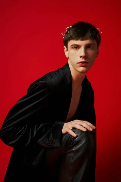 Hombre guapo en traje negro con pantalones de cuero y flores en el pelo sentado sobre fondo rojo - foto de stock