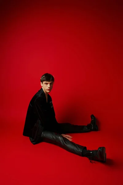 Красивый молодой человек в черной одежде с цветущими цветами в волосах, сидящих на красном фоне — стоковое фото