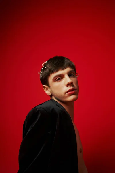 Junger modischer und hemdloser Mann im Samtblazer mit Blumen im Haar posiert auf rotem Hintergrund — Stockfoto