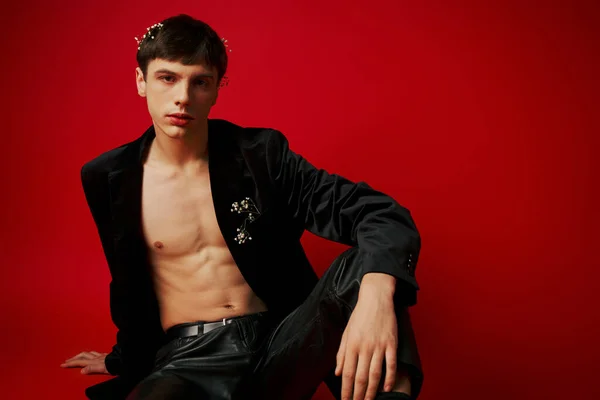 Young male model in black attire with flowers in hair and leather pants sitting on red background — Stock Photo