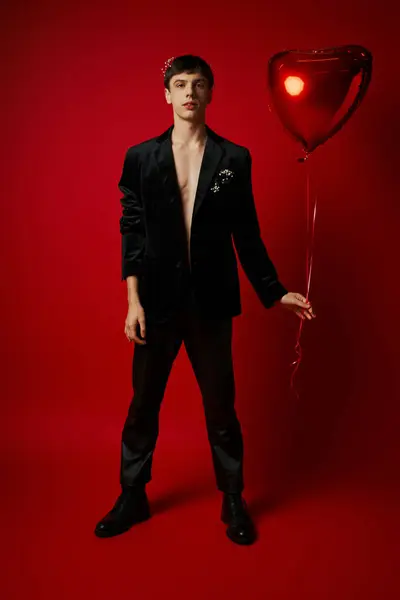 Full length of handsome young gentleman in black attire holding heart-shaped balloon on red backdrop — Stock Photo