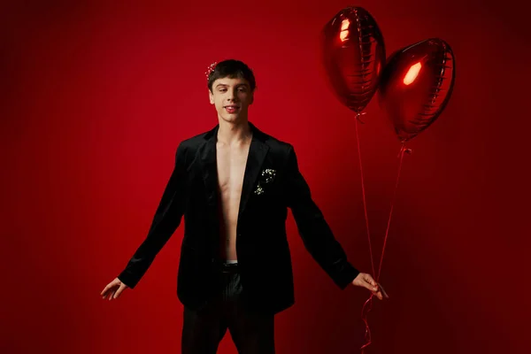 Jovem feliz e bonito em traje preto segurando balões em forma de coração no fundo vermelho — Fotografia de Stock