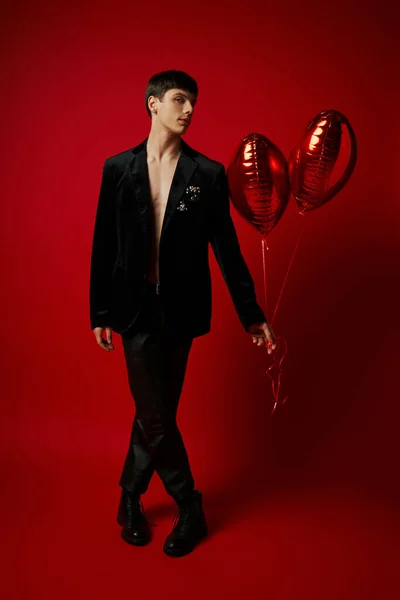 Comprimento total de cavalheiro elegante em traje preto segurando balões em forma de coração no fundo vermelho — Fotografia de Stock
