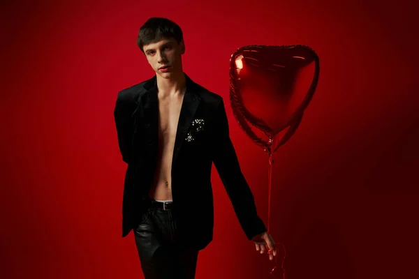 Élégant modèle masculin en veste de velours et pantalon en cuir tenant des ballons en forme de coeur sur fond rouge — Photo de stock