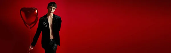 Стильний чоловік в оксамитовій куртці і шкіряні штани, що тримають повітряні кулі у формі серця на червоному тлі, банер — стокове фото