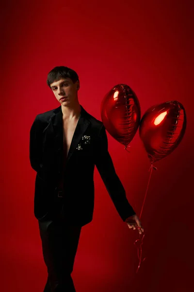 Jovem modelo masculino em jaqueta de veludo e calças de couro segurando balões em forma de coração no fundo vermelho — Fotografia de Stock