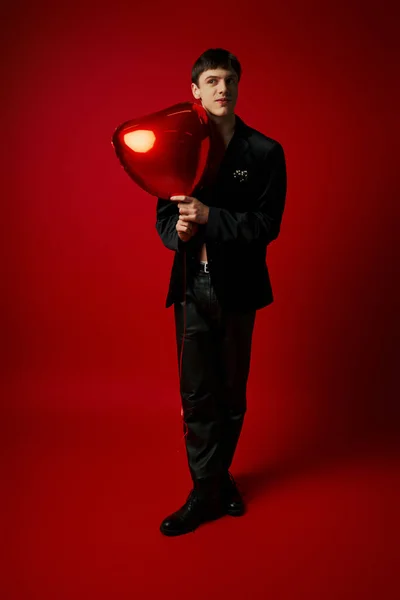 Happy male model in velvet jacket and leather pants holding heart-shaped balloon on red backdrop — Stock Photo