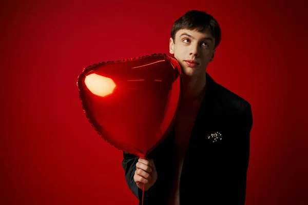 Jovem confuso em casaco de veludo segurando balão em forma de coração e olhando para o fundo vermelho — Fotografia de Stock