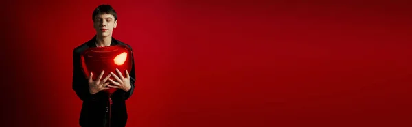 Happy gen z guy in velvet jacket smiling and holding heart-shaped balloon on red background, banner — Stock Photo