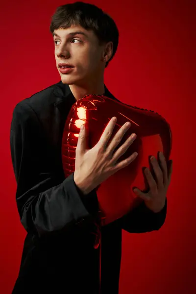 Handsome gen z guy in velvet jacket smiling and holding heart-shaped balloon on red background — Stock Photo