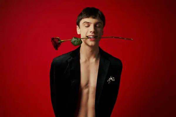 Красивий молодий чоловік в оксамитовій куртці тримає троянду в зубах і посміхається на червоному тлі, романтика — стокове фото