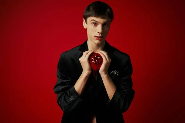 Romántico chico sosteniendo en forma de corazón presente y mirando a la cámara en el fondo rojo, día de San Valentín - foto de stock