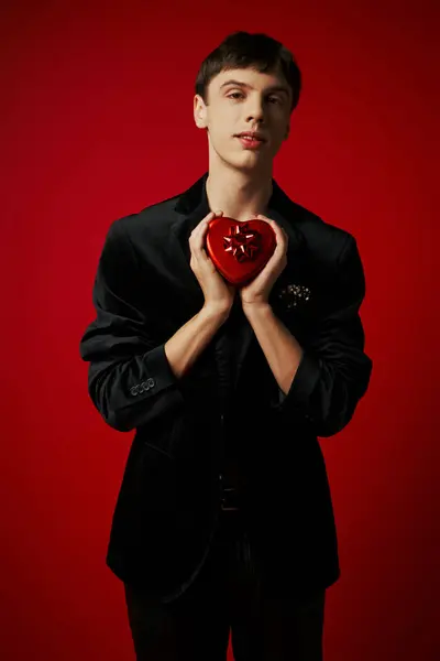 Romántico hombre sosteniendo en forma de corazón presente y mirando a la cámara en el fondo rojo, día de San Valentín - foto de stock