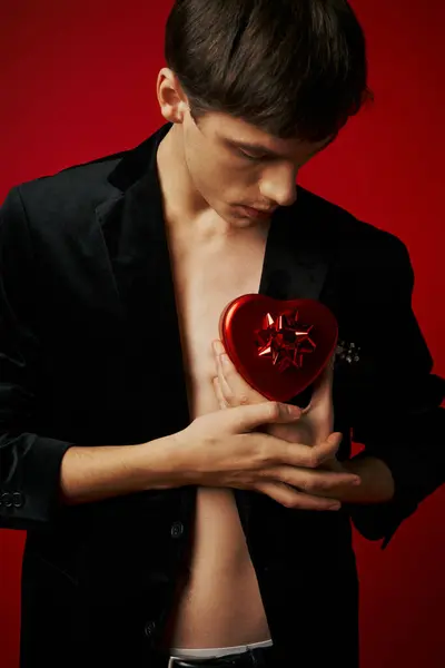 Giovane uomo romantico in velluto blazer tenendo regalo a forma di cuore su sfondo rosso, San Valentino — Foto stock