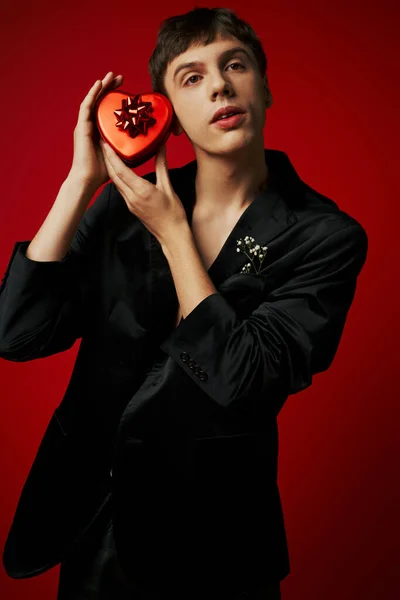 Bell'uomo giovane in velluto blazer che tiene regalo a forma di cuore su sfondo rosso, San Valentino — Foto stock