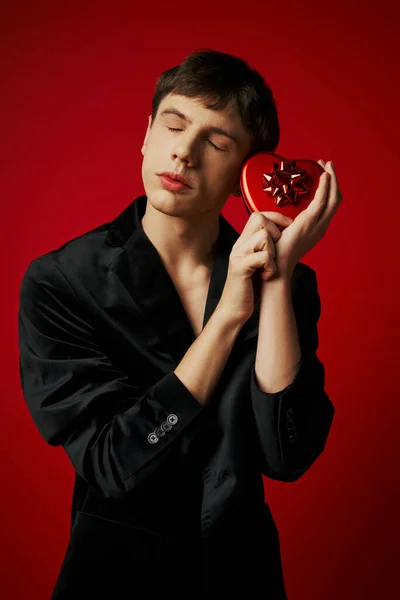 Sentimental man in velvet blazer holding heart-shaped present on red background, Valentines day — Stock Photo