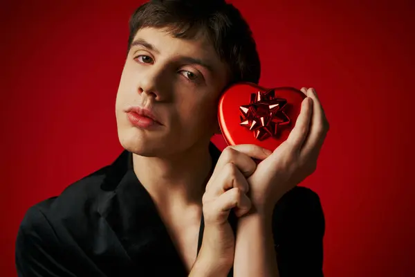Homme sentimental en velours blazer tenant boîte cadeau en forme de coeur sur fond rouge, Saint Valentin — Photo de stock