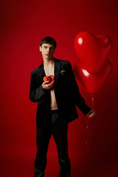 Romántico joven sosteniendo caja de regalo en forma de corazón y globos sobre fondo rojo, día de San Valentín - foto de stock