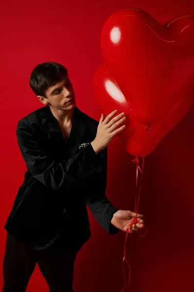 Romantic young man in velvet attire holding heart-shaped balloons on red background, Valentines day — Stock Photo