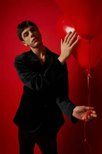 Romantique jeune homme en tenue noire tenant des ballons en forme de coeur sur fond rouge, Saint Valentin — Photo de stock
