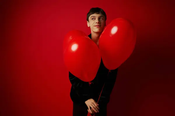 Joven feliz en traje negro sosteniendo globos en forma de corazón sobre fondo rojo, día de San Valentín - foto de stock