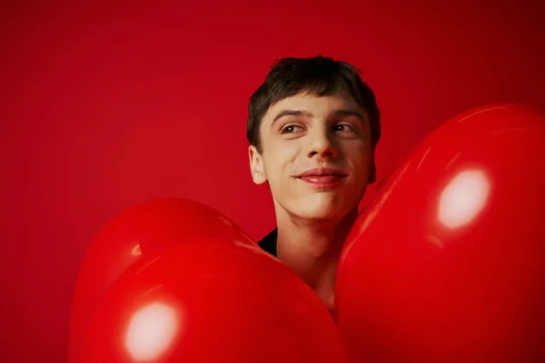 Feliz joven sonriendo alrededor de globos en forma de corazón sobre fondo rojo, concepto del día de San Valentín - foto de stock