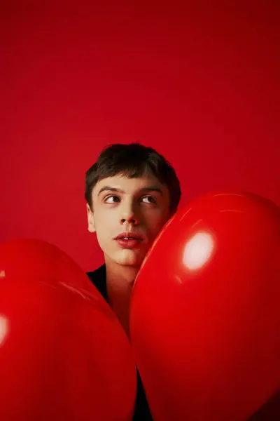 Romántico y soñador hombre cerca de globos en forma de corazón sobre fondo rojo, concepto del día de San Valentín - foto de stock
