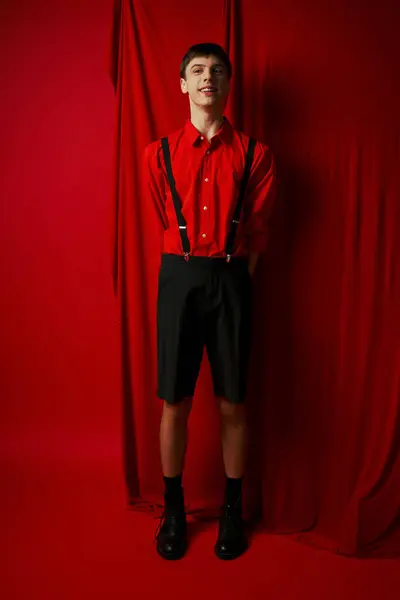 Happy young man in shorts with suspenders and shirt standing near red curtain, fashionable look — Stock Photo
