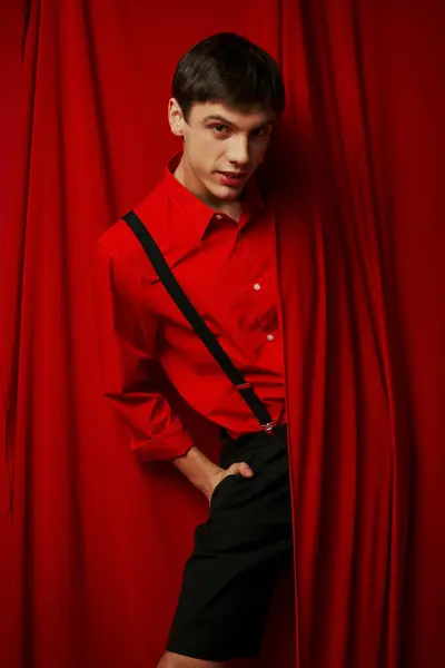 Glücklicher junger Mann in lebendigem Hemd mit Hosenträgern, die sich hinter rotem Vorhang verstecken, modischer Look — Stockfoto