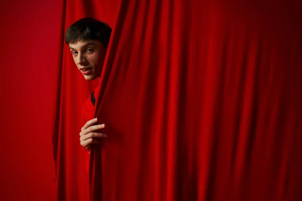 Jovem brincalhão em camisa vibrante escondendo atrás de cortina vermelha enquanto joga esconde e procura, espreitando — Fotografia de Stock
