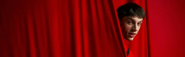 Playful young man in vibrant shirt hiding behind red curtain while playing hide and seek, banner — Stock Photo