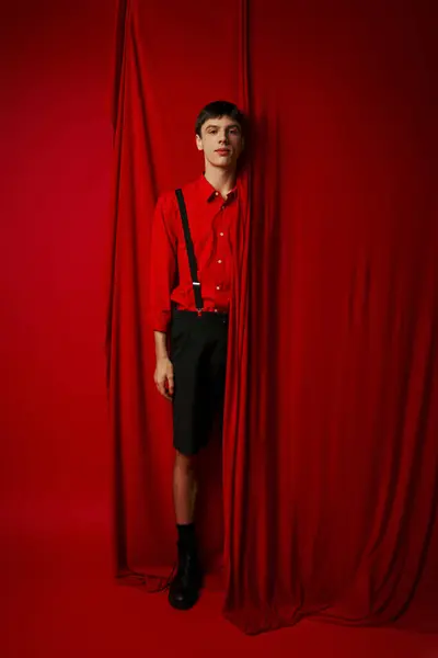 Full length of young man in vibrant shirt with suspenders hiding behind red curtain, trendy look — Stock Photo