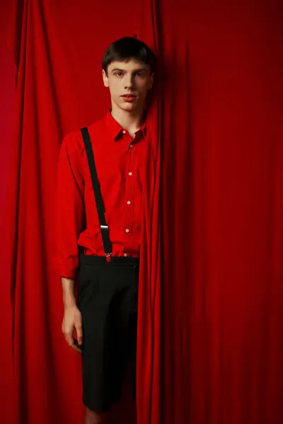 Young man in vibrant shirt and shorts with suspenders hiding behind red curtain, trendy look — Stock Photo