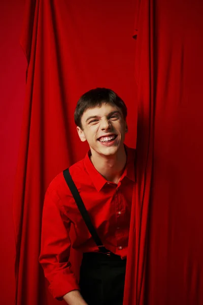 Joyeux jeune homme en jarretelles souriant tout en se cachant derrière un rideau rouge vibrant, gaieté — Photo de stock