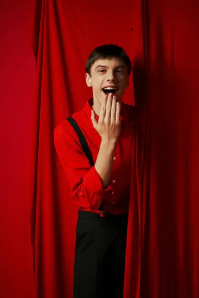 Здивований молодий чоловік у підтяжках посміхається і покриває рот біля червоної яскравої завіси, веселощі — стокове фото