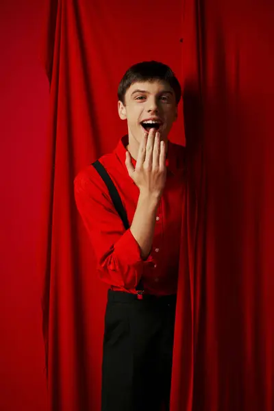 Jeune homme étonné dans les bretelles souriant et couvrant la bouche près rouge rideau vibrant, gaieté — Photo de stock