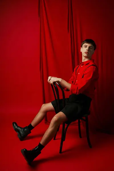 Hombre joven en camisa y pantalones cortos negros sentado en silla con pose segura al lado de la cortina roja - foto de stock