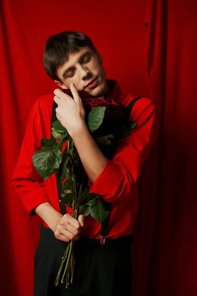 Tranquilo y joven hombre en pantalones cortos con tirantes abrazando ramo de rosas rojas cerca de la cortina - foto de stock