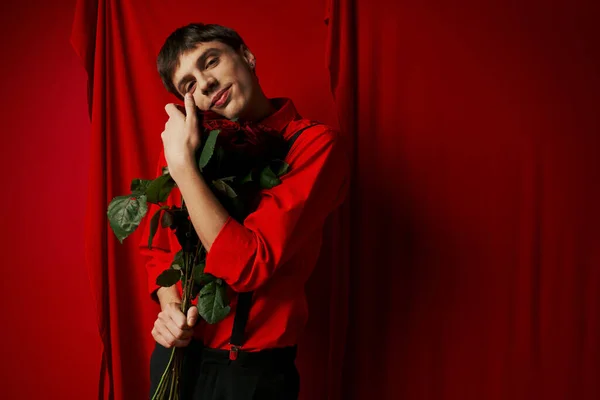 Joven contento en pantalones cortos con tirantes abrazando ramo de rosas rojas cerca de cortina vibrante - foto de stock