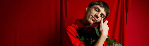 Heureux jeune homme en short avec bretelles embrassant bouquet de roses près rideau rouge, bannière — Photo de stock
