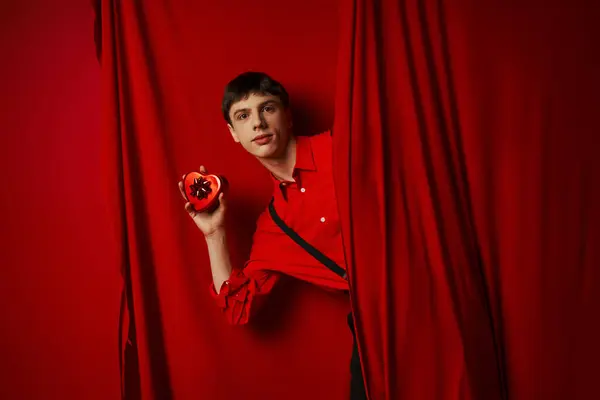 Joven de camisa roja con tirantes sosteniendo caja de regalo en forma de corazón cerca de la cortina, 14 de febrero - foto de stock