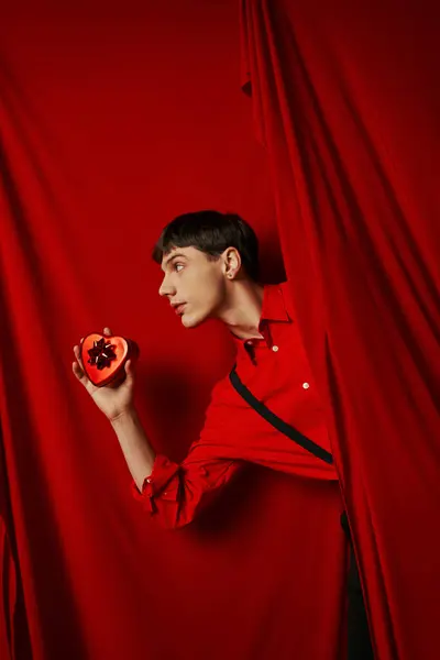 Cara em camisa vermelha com suspensórios segurando caixa de presente em forma de coração perto de cortina, dia dos namorados — Fotografia de Stock