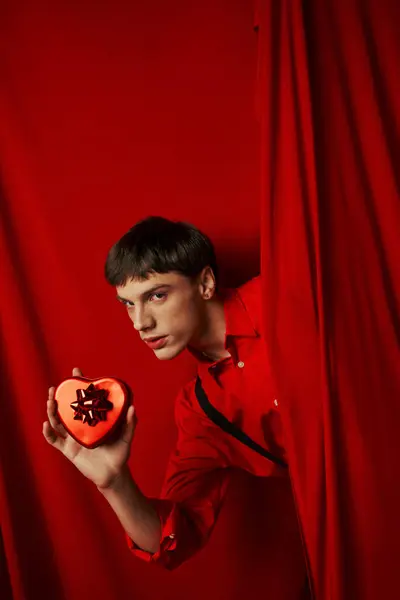 Giovane uomo in camicia rossa con bretelle in possesso di cuore a forma di regalo vicino alla tenda, San Valentino — Foto stock