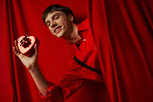 Felice uomo in camicia rossa con bretelle in possesso di cuore a forma di regalo vicino alla tenda, San Valentino — Foto stock