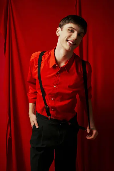 Cheerful young man in shirt and suspenders posing with hand in pocket of pants on red background — Stock Photo