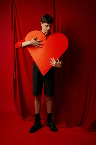 Romantic young man in black shorts embracing large heart cutout on red background, Valentines day — Stock Photo
